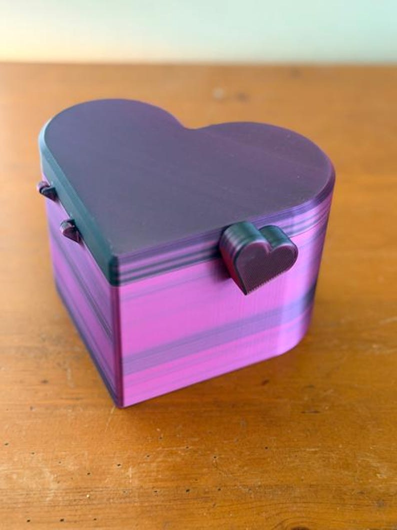 May include: A heart-shaped box with a hinged lid, featuring a gradient of dark purple and vibrant magenta horizontal stripes. The lid includes a small, heart-shaped latch on the front, and the entire structure displays visible 3D printed layers.