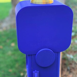 May include: A blue 3D-printed gumball machine featuring a square-shaped container, a gold-coloured circular lid, a round dispensing knob on the front, and a small blue lever at the base. The device has a smooth matte finish and a boxy structure.