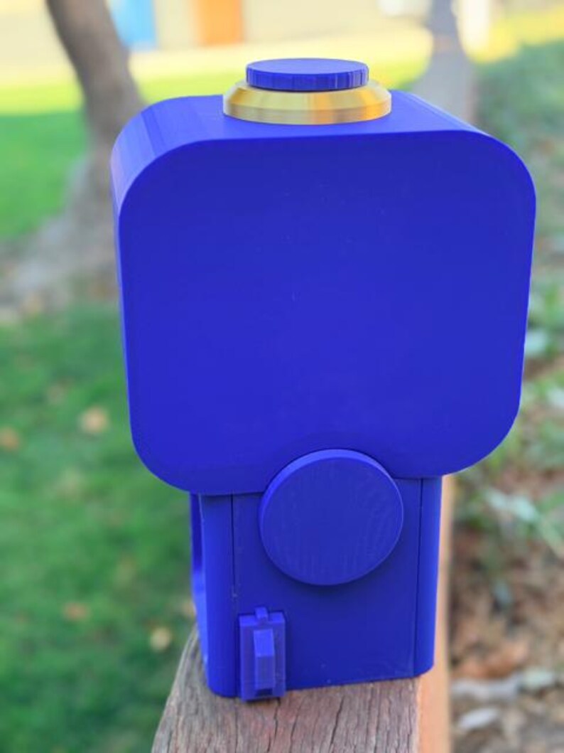 May include: A blue 3D-printed gumball machine featuring a square-shaped container, a gold-coloured circular lid, a round dispensing knob on the front, and a small blue lever at the base. The device has a smooth matte finish and a boxy structure.