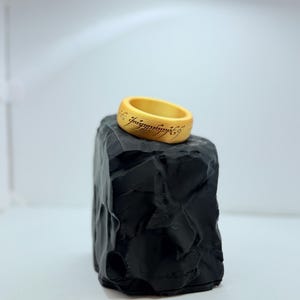 以下が含まれることがあります： 外側の表面に黒い刻印文字が施された、滑らかで丸みを帯びたゴールドカラーのリング。リングは、暗い色でギザギザとした岩のような質感のディスプレイスタンドの上に置かれています。