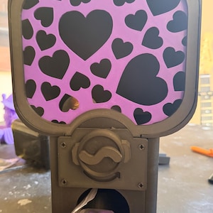 May include: A black gumball-style dispenser featuring a purple panel decorated with black hearts. The device has a central rotating knob and a dispensing chute at the bottom, which contains a small clear plastic bag filled with purple circular tokens.