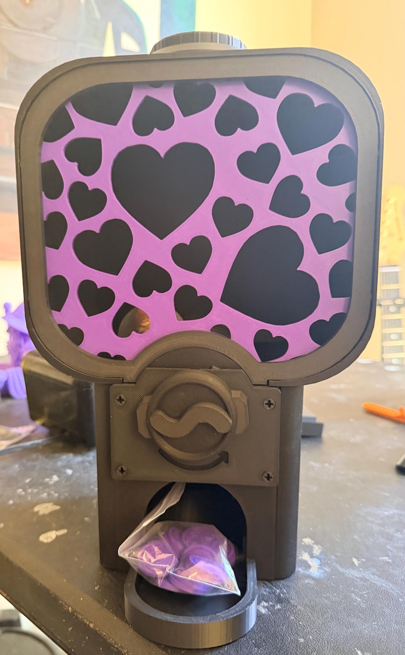 May include: A black gumball-style dispenser featuring a purple panel decorated with black hearts. The device has a central rotating knob and a dispensing chute at the bottom, which contains a small clear plastic bag filled with purple circular tokens.
