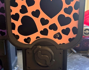 May include: A black vending machine with a rectangular front panel featuring a repeating pattern of black hearts on an orange background. The machine has a central black dial and a dispensing slot containing a clear plastic bag of orange tokens.