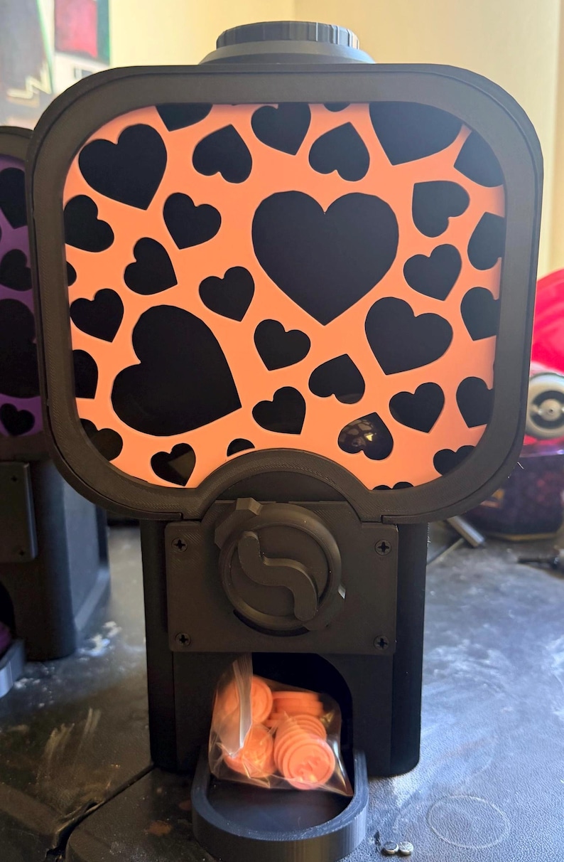 May include: A black vending machine with a rectangular front panel featuring a repeating pattern of black hearts on an orange background. The machine has a central black dial and a dispensing slot containing a clear plastic bag of orange tokens.