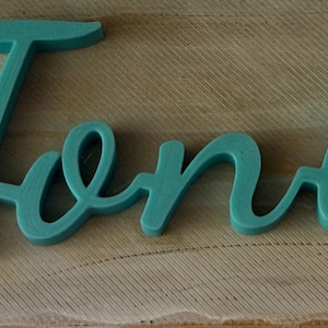 May include: A teal-colored, three-dimensional script sign featuring the name Toni in a cursive font. The letters are connected in a single piece, displaying a smooth, matte finish with rounded edges, creating a distinct and legible decorative word.