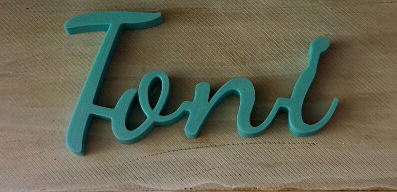 May include: A teal-colored, three-dimensional script sign featuring the name Toni in a cursive font. The letters are connected in a single piece, displaying a smooth, matte finish with rounded edges, creating a distinct and legible decorative word.