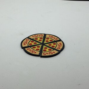 May include: A circular game token shaped like a pizza, divided into six individual triangular slices. Each slice features a yellow crust, red pepperoni dots, and the text +5/+5 printed in red. The entire token has a black border.