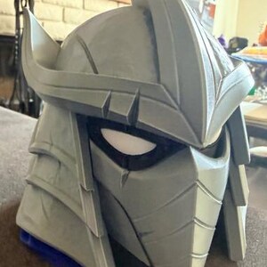 May include: A gray, angular helmet featuring a prominent central crest, layered side panels, and a face mask with narrow, dark eye openings. The design includes sharp, geometric lines and a pointed top section, creating a structured, protective appearance.