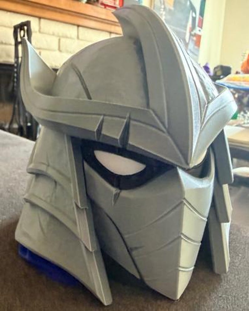 May include: A gray, angular helmet featuring a prominent central crest, layered side panels, and a face mask with narrow, dark eye openings. The design includes sharp, geometric lines and a pointed top section, creating a structured, protective appearance.