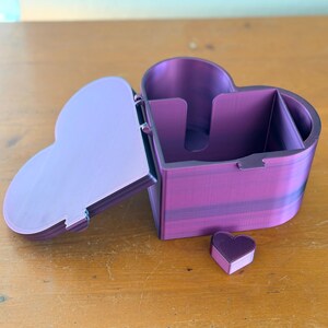 May include: A heart-shaped storage box with a hinged lid, featuring a vibrant purple and magenta gradient finish. The interior is divided into sections, and a small, separate heart-shaped piece sits in front of the main container.