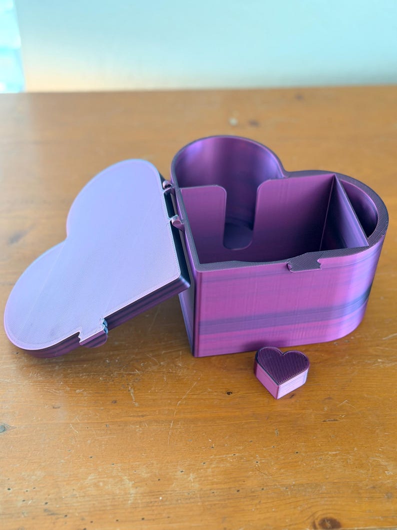 May include: A heart-shaped storage box with a hinged lid, featuring a vibrant purple and magenta gradient finish. The interior is divided into sections, and a small, separate heart-shaped piece sits in front of the main container.