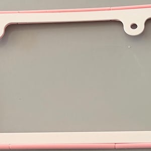 May include: A light pink and white number plate surround with rounded edges. The left side features the text "Scan Me" in raised pink lettering, while the right side has a raised pink square. The frame includes two circular mounting holes at the top.