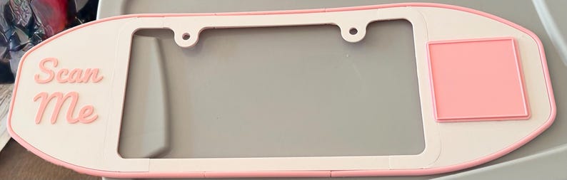 May include: A light pink and white number plate surround with rounded edges. The left side features the text "Scan Me" in raised pink lettering, while the right side has a raised pink square. The frame includes two circular mounting holes at the top.