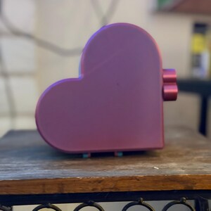 May include: A heart-shaped object with a smooth, matte purple finish. The side of the object features two small, cylindrical, protruding buttons or knobs in a matching purple color. The overall form is rounded and symmetrical in a heart silhouette.