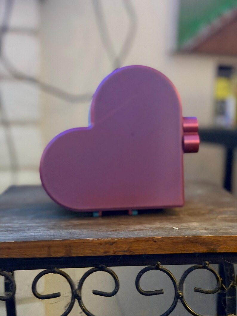 May include: A heart-shaped object with a smooth, matte purple finish. The side of the object features two small, cylindrical, protruding buttons or knobs in a matching purple color. The overall form is rounded and symmetrical in a heart silhouette.