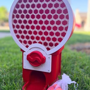 May include: A red and white gumball machine featuring a circular top section with a honeycomb pattern. The machine has a central red dispensing knob on a white square plate and a bottom chute. A small white organza bag filled with red tokens sits nearby.