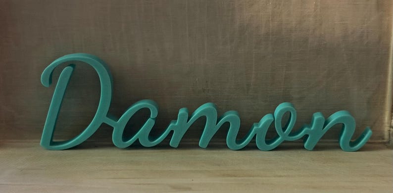 May include: A teal-colored, three-dimensional script sign displaying the name Damon in a continuous, flowing cursive font. The letters are connected and feature a smooth, matte finish, standing upright as a decorative piece of lettering.