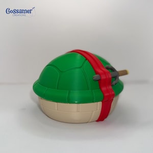 May include: A green turtle shell with a red headband and a weapon. The shell has a beige base and a green, segmented top. The red headband wraps around the shell. A silver and brown weapon is attached.