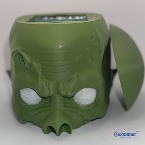 May include: A green, stylized goblin head with a hinged lid. The goblin has white eyes, a prominent nose, and textured skin. The product is likely a container or a decorative item.