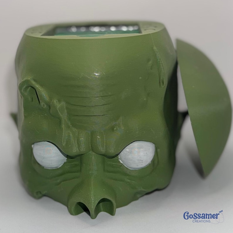 May include: A green, stylized goblin head with a hinged lid. The goblin has white eyes, a prominent nose, and textured skin. The product is likely a container or a decorative item.