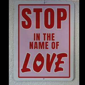 May include: A pink and red sign with the text "STOP IN THE NAME OF LOVE" in bold white letters. The word "LOVE" is in a stylized font with a small white heart in the center.