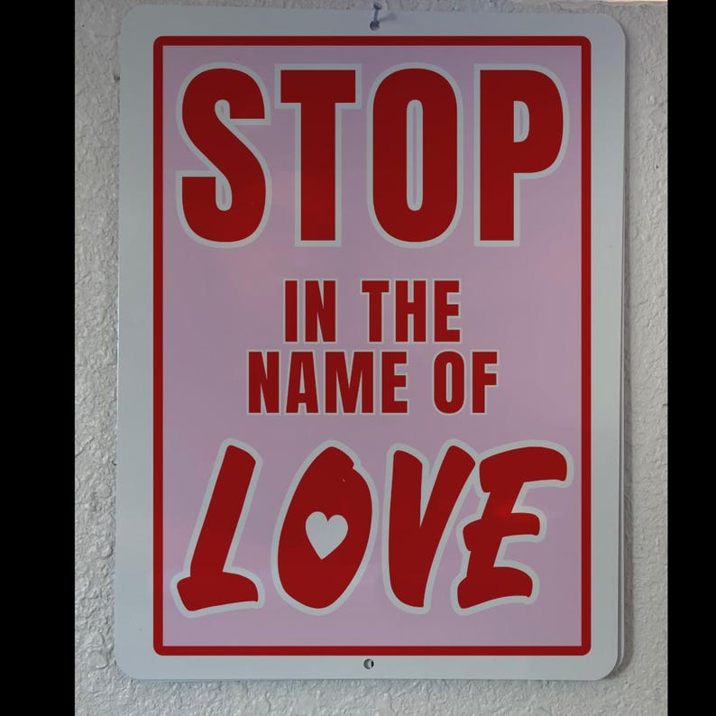May include: A pink and red sign with the text "STOP IN THE NAME OF LOVE" in bold white letters. The word "LOVE" is in a stylized font with a small white heart in the center.