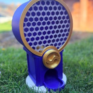 May include: A blue and gold gumball machine featuring a circular container with a honeycomb pattern. The device has a gold-colored dispensing knob, a blue base, and a small chute at the bottom for dispensing items. The structure is upright and cylindrical.