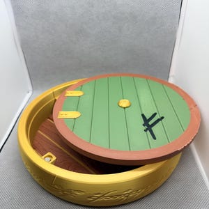 May include: A round yellow container with a removable green circular lid featuring a central gold knob, two gold hinges, and a black symbol. The yellow base has an embossed script pattern around the exterior, and the interior floor is brown.