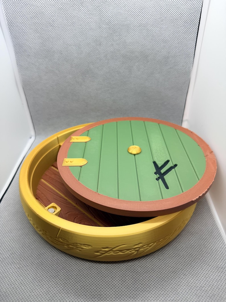 May include: A round yellow container with a removable green circular lid featuring a central gold knob, two gold hinges, and a black symbol. The yellow base has an embossed script pattern around the exterior, and the interior floor is brown.