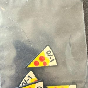 May include: Four triangular pizza slice charms with yellow cheese, red pepperoni circles, and a cream-colored crust edge. Each slice features the black text 0/1 printed on the crust. The charms are contained within a clear plastic zip-top bag.