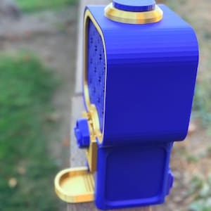 May include: A blue and gold dispenser featuring a rectangular body with a rounded top, a circular button on the upper surface, a front-facing panel with a grid pattern, a central knob, and a small protruding tray at the base.