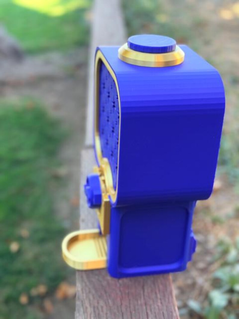 May include: A blue and gold dispenser featuring a rectangular body with a rounded top, a circular button on the upper surface, a front-facing panel with a grid pattern, a central knob, and a small protruding tray at the base.