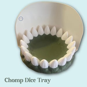 Puede incluir: Una bandeja de dados novedosa con forma de boca, con dientes blancos alrededor de un interior verde. La bandeja tiene una parte posterior blanca y curvada y el texto "Chomp Dice Tray" en la parte inferior.