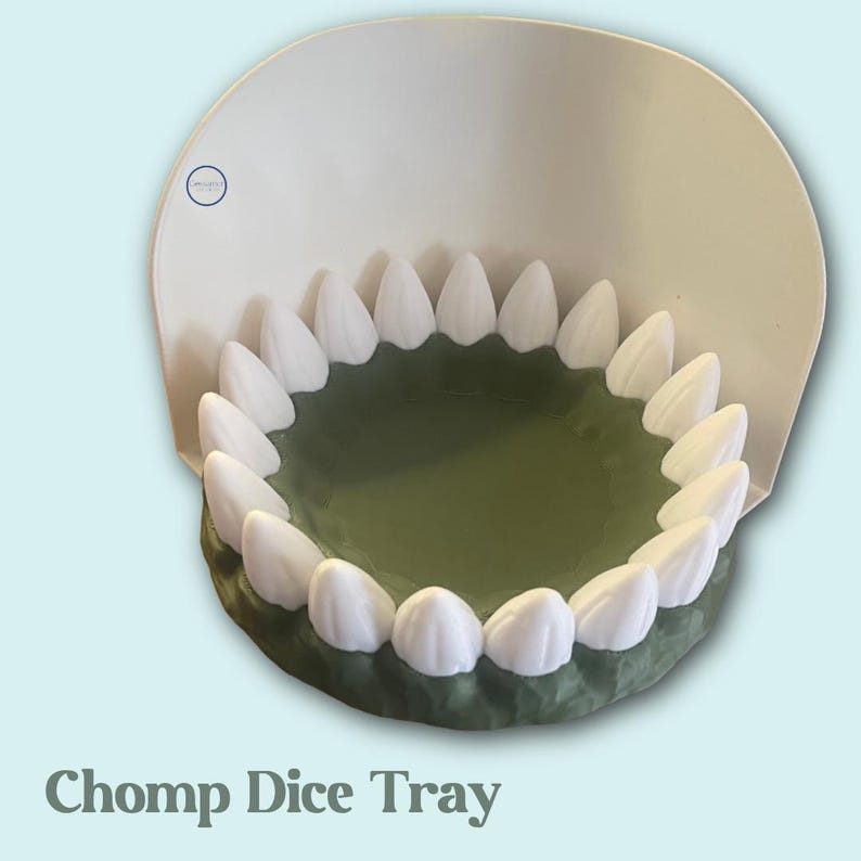 Puede incluir: Una bandeja de dados novedosa con forma de boca, con dientes blancos alrededor de un interior verde. La bandeja tiene una parte posterior blanca y curvada y el texto "Chomp Dice Tray" en la parte inferior.