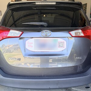 May include: A grey Toyota RAV4 XLE vehicle rear view featuring a number plate frame with a pink border, the text Scan Me in pink script, and a square QR code on the right side. The car has a rear windscreen wiper and a chrome Toyota emblem.