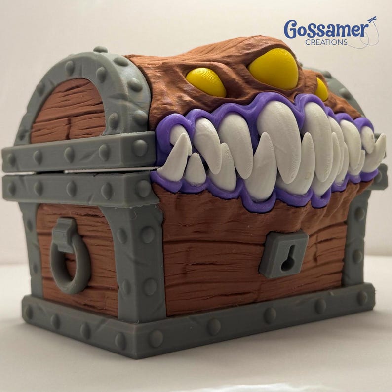 May include: A brown treasure chest with a monster face design. The chest features a hinged lid, gray metal accents, and a keyhole. The monster face has yellow eyes, large white teeth, and purple gums. The chest is labeled "Gossamer Creations".
