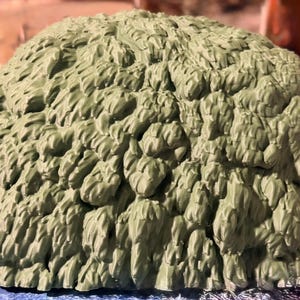 May include: A dome-shaped object with a textured, sage green surface featuring numerous overlapping, leaf-like or brushstroke-style protrusions that create a dense, layered appearance across the entire rounded exterior of the item.