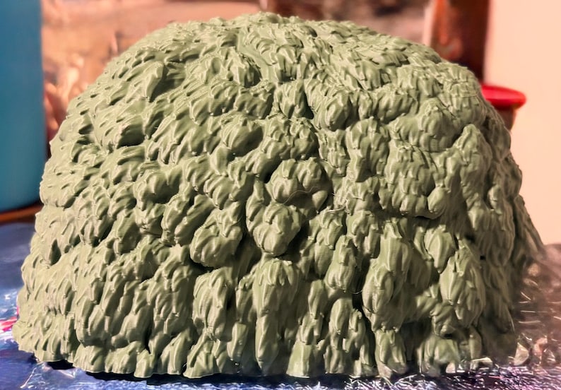May include: A dome-shaped object with a textured, sage green surface featuring numerous overlapping, leaf-like or brushstroke-style protrusions that create a dense, layered appearance across the entire rounded exterior of the item.