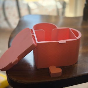 May include: A light pink heart-shaped storage box with a hinged lid that is currently open. The interior of the box is divided into two sections. A small, separate heart-shaped piece is placed on the surface in front of the main container.