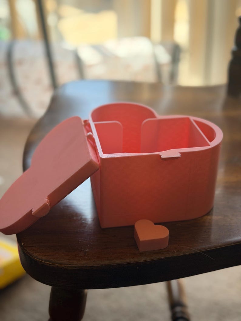 May include: A light pink heart-shaped storage box with a hinged lid that is currently open. The interior of the box is divided into two sections. A small, separate heart-shaped piece is placed on the surface in front of the main container.