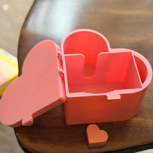 May include: A heart-shaped storage box in a solid light pink color with a hinged lid that is currently open. The interior features a divided compartment structure. A small, separate heart-shaped piece in a matching pink tone sits in front.