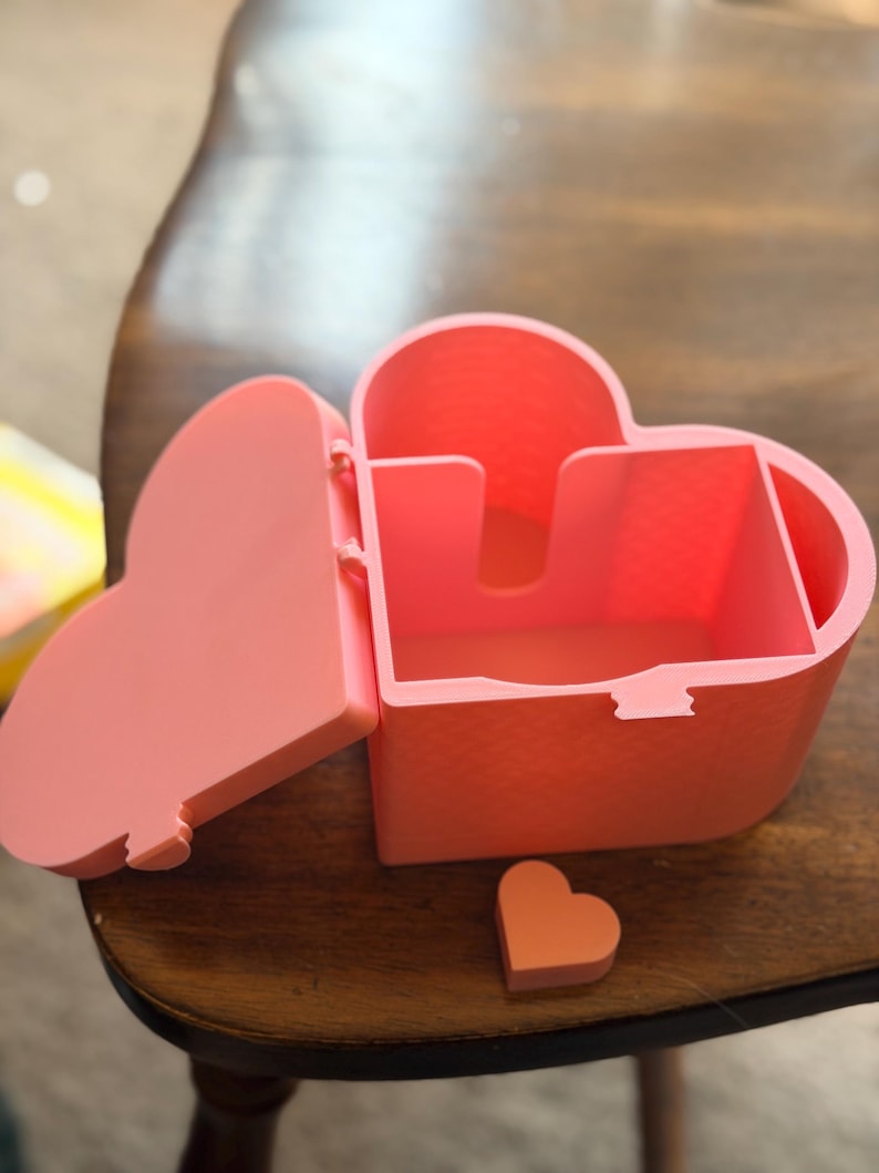 May include: A heart-shaped storage box in a solid light pink color with a hinged lid that is currently open. The interior features a divided compartment structure. A small, separate heart-shaped piece in a matching pink tone sits in front.