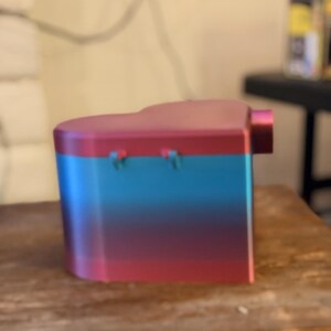 May include: A heart-shaped container featuring a gradient color scheme that transitions from a vibrant red at the top and bottom to a bright blue in the center. The object has a smooth, solid surface with two small hinges visible on the side.