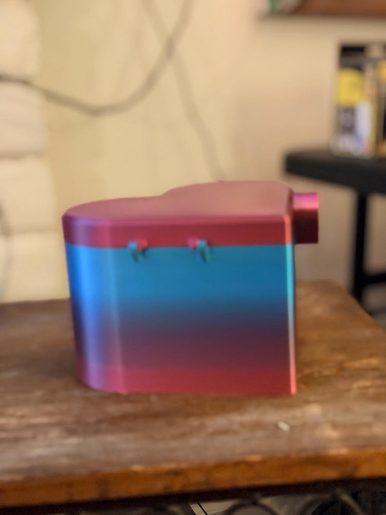 May include: A heart-shaped container featuring a gradient color scheme that transitions from a vibrant red at the top and bottom to a bright blue in the center. The object has a smooth, solid surface with two small hinges visible on the side.