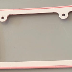 May include: A light pink and white license plate frame with rounded edges. The left side features the text "Scan Me" in raised pink lettering, while the right side has a raised pink square. The frame includes two circular mounting holes at the top.