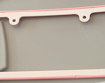 May include: A light pink and white license plate frame with rounded edges. The left side features the text "Scan Me" in raised pink lettering, while the right side has a raised pink square. The frame includes two circular mounting holes at the top.