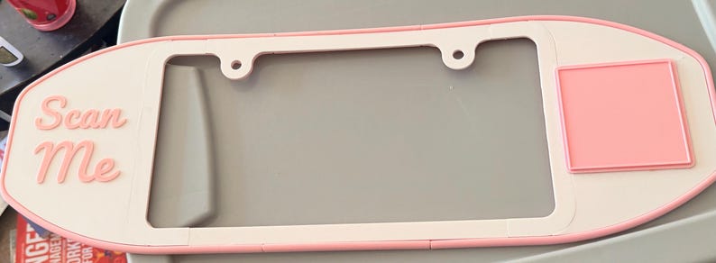 May include: A light pink and white number plate surround with rounded edges. The left side features the text "Scan Me" in raised pink lettering, while the right side has a raised pink square. The frame includes two circular mounting holes at the top.