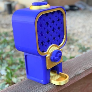 May include: A blue and gold gumball machine featuring a square top section with a star-patterned front panel, a circular lid on top, and a gold dispensing tray at the base. The device has a central gold mechanism with a blue knob for operation.