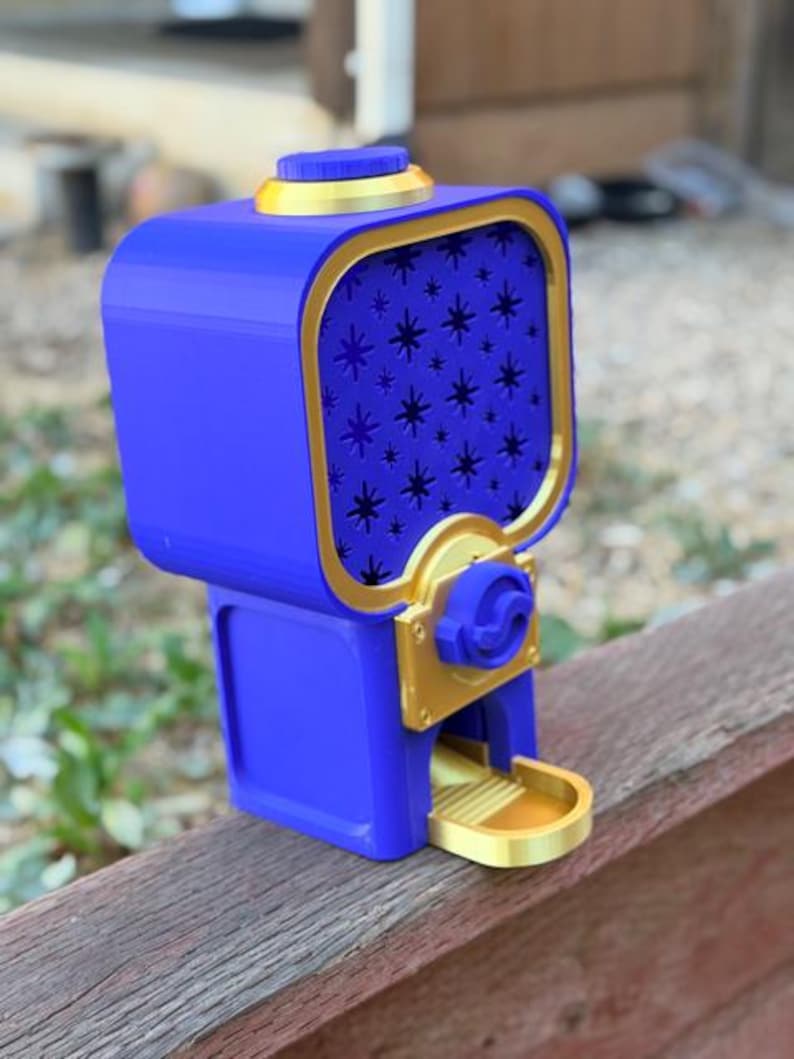 May include: A blue and gold gumball machine featuring a square top section with a star-patterned front panel, a circular lid on top, and a gold dispensing tray at the base. The device has a central gold mechanism with a blue knob for operation.