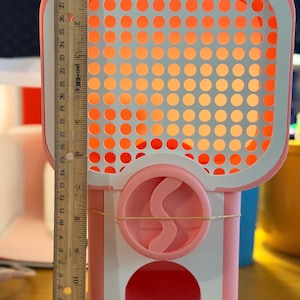 May include: A pink and white plastic gumball machine featuring a grid-patterned front panel and a circular dispensing mechanism. A wooden ruler is placed vertically along the left side, showing the machine stands approximately 29 cm tall.